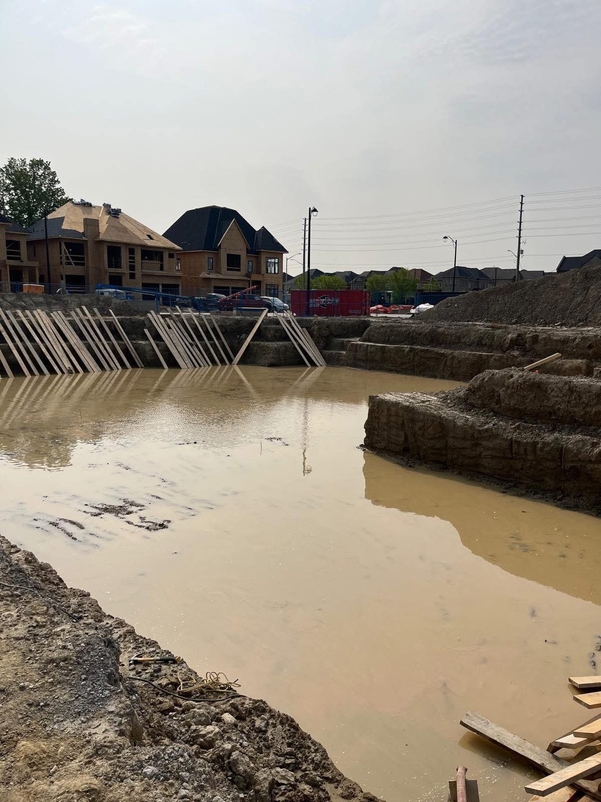 Construction site with flooded excavation pit, metal drainage barriers, and residential buildings in background — MAY Engineering construction management Ontario tendering and bid review